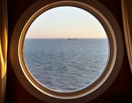 Blick durch ein rundes Bullauge auf das Meer bei Sonnenuntergang, in der Ferne ist ein Schiff zu sehen.