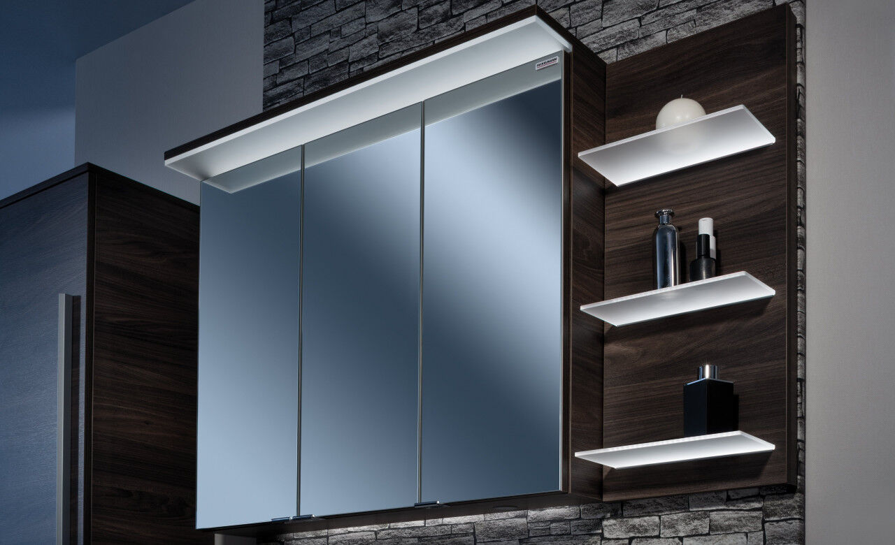 Spiegelschrank mit LED-Beleuchtung in einem dunklen Bad, neben dem Spiegelschrank leuchten zusätzliche Regelböden weiß.