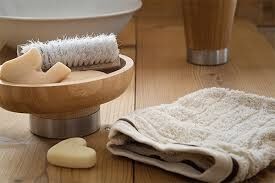 Auf einem braunen Holzboden steht eine hölzerne Schalen mit Seifen und einer Bürste. Rechts daneben liegt ein beiger Waschlappen, links davon eine Seife in Herzform.